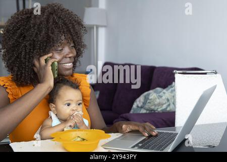 Cheerful African mother using a laptop while feeding her baby girl and talking on a phone at home Stock Photo