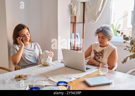 A mature woman works on a laptop while holding a small dog, as her young adult daughter chats on a mobile phone during breakfast Stock Photo