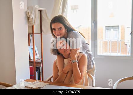 A touching scene of a young adult daughter embracing her smiling middle-aged mother from behind, both enjoying a serene moment together in a brightly Stock Photo