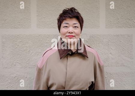 A smiling mature Asian woman stands confidently in front of a textured beige wall Her stylish multicolored coat with a brown collar adds to her cheerf Stock Photo