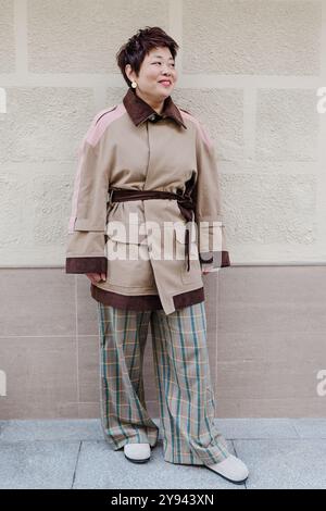 Middle-aged Asian woman dressed in a fashionable trench coat with pink accents and plaid pants, smiling confidently against a light wall background Stock Photo
