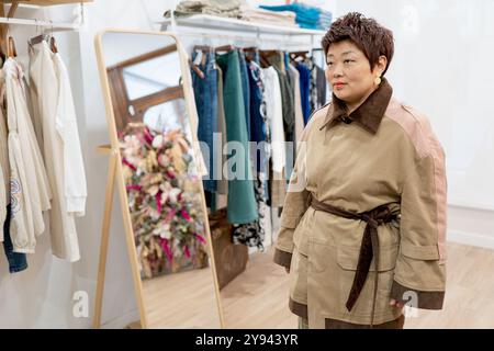 An Asian woman wearing an elegant brown and beige trench coat stands thoughtfully in a fashionable clothing store, surrounded by a variety of fashion Stock Photo