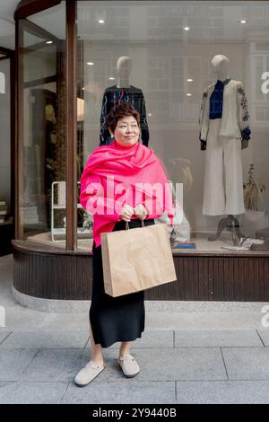 A joyful mature asian woman in a pink scarf stands in front of a clothes store, carrying a shopping bag and wearing stylish attire Stock Photo