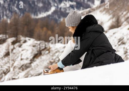 Side view of an Asian woman adjusting her snowshoes while sitting in snow-covered mountain terrain. She wears a black jacket and gray beanie, calmly s Stock Photo