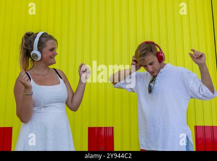 A young woman and a young man stand against a vibrant yellow wall, both wearing headphones and dancing to their own rhythm under a clear sky Stock Photo