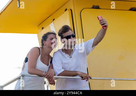 A young couple enjoys a sunny day at the beach in autumn, taking a selfie together, with a vibrant yellow structure in the background, highlighting a Stock Photo