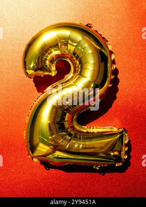 A shiny golden number two balloon brightly lit and centered against a sparkling red glitter background, reflecting a sense of celebration. Stock Photo