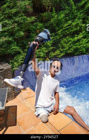 A happy, middle-aged man with a prosthetic leg is sitting by the edge of a pool, playfully lifting his prosthetic limb with a smile Stock Photo