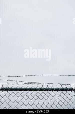 Close-up view of a chain-link fence topped with barbed wire against a grey, cloudy sky. This urban scene captures the sense of isolation and security Stock Photo