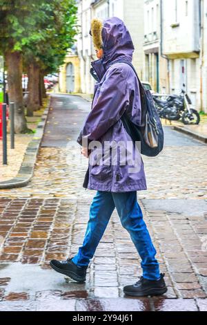 Woman with rainwear walking along city street in pouring rain without umbrella - Tours, Indre-et-Loire (37), France. Stock Photo