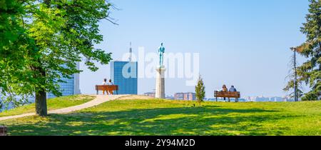 Visitors relax on benches near the Victor Monument, overlooking the expansive views from Kalemegdan Fortress, a historic site in Belgrade, Serbia, under a clear blue sky. Stock Photo