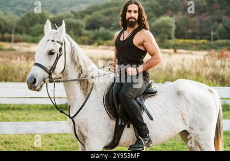 Young jockey training his horse for a ride. Good looking, hunky cowboy rides horse at beautiful country side nature. Equestrian rides his white well Stock Photo