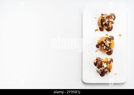 Profiteroles or eclairs poured with chocolate glaze with almond nut petals on a white plate and a light background. View from above. Space for text. C Stock Photo