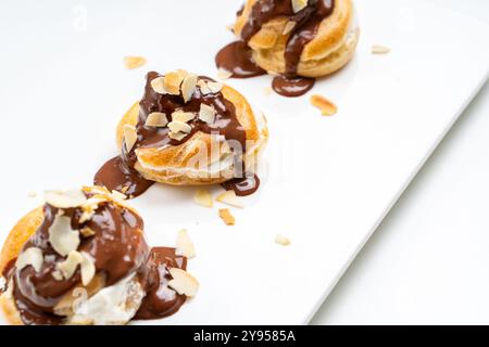 Profiteroles or eclairs poured with chocolate glaze with almond nut petals on a white plate and a light background. View from above. Space for text. C Stock Photo