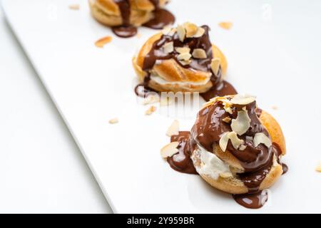 Profiteroles or eclairs poured with chocolate glaze with almond nut petals on a white plate and a light background. View from above. Space for text. C Stock Photo