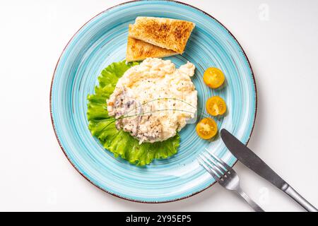 Scrambled eggs with mushrooms and crispy toast garnished with lettuce and yellow cherry tomatoes, on a turquoise plate, on a white table with cutlery. Stock Photo