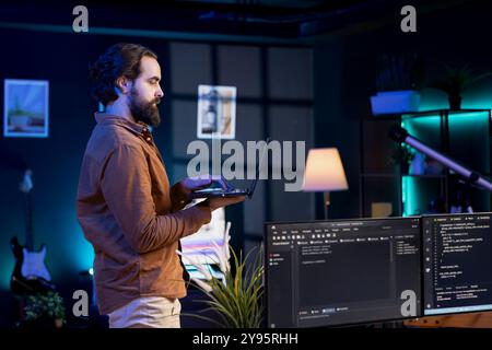 Developer doing software debugging using laptop, checking glitches in lines of code displayed on PC monitors. Man walking around home fixing coding issues using notebook device Stock Photo