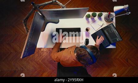 Top down shot of technician focused on typing complex code in home office, developing software application. It specialist removing headphones to better concentrate on difficult task, aerial shot Stock Photo