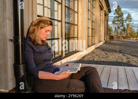 Beautiful woman enjoys reading a book while sitting against the wall on a wooden deck, with a modern cabin and trees during the summer Stock Photo