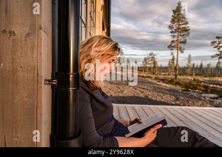 A beautiful woman sits peacefully on a deck, reading a book with mountains and trees in the background during golden hour, embracing nature Stock Photo