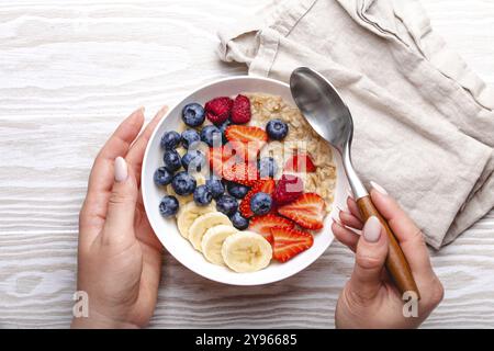 Top view of female hands holding bowl with oatmeal porridge with fruit and berries and spoon on white wooden background table top view, woman eating h Stock Photo