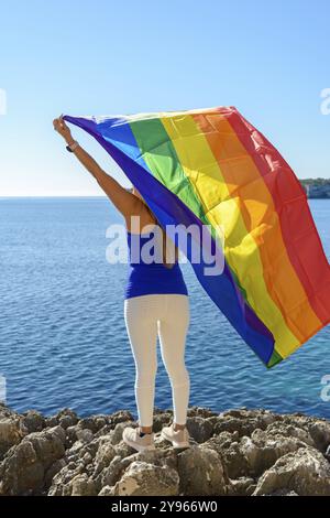 A woman in active wear holds a rainbow flag facing the ocean on rocks, Woman, middle aged, outdoors waving pride flag, concept of libertat, equality. Stock Photo