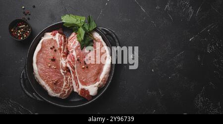 Cut raw meat pork steaks with seasonings in black cast iron pan, dark rustic stone background top view, ready for roasting. Pork loin chops cooking wi Stock Photo