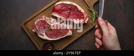 Male's hand holding knife over two raw uncooked meat beef rib eye marbled steaks on wooden cutting board and seasonings on dark rustic background from Stock Photo