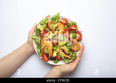 Female hands holding healthy salad with grilled shrimps, avocado, cherry tomatoes and green leaves on white plate isolated on white background top vie Stock Photo