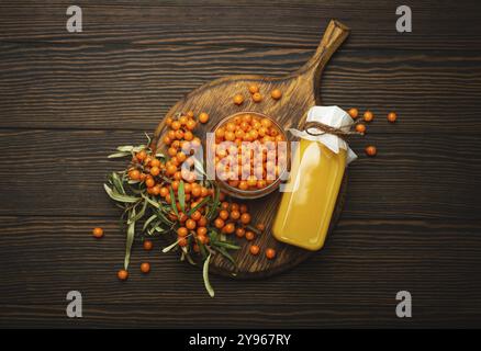 Sea buckthorn healthy drink in bottle, ripe berries in glass jar and branches with leaves top view on dark wooden rustic background, great for skin, h Stock Photo