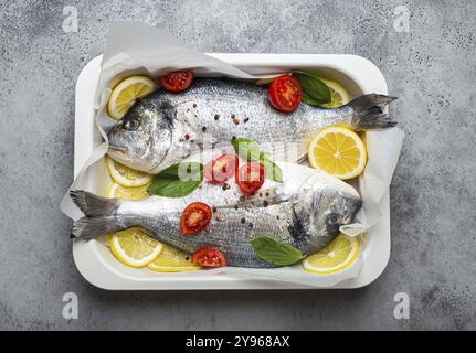 Two raw fishes dorado in casserole dish with ingredients lemon, fresh basil, cherry tomatoes close up top view on rustic stone grey background, cookin Stock Photo