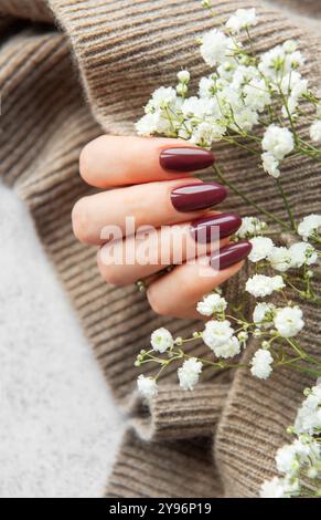 A hand with long, deep burgundy nails gently holds a sprig of small white flowers against a soft striped sweater, blending beauty and comfort. Stock Photo