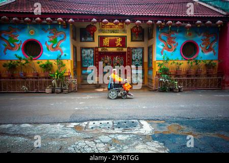 A monk on a wheelchair is collecting alms passes by a Chinese shrine that was flooded, with clear water stains visible at the door. After the flood situation in Chiang Mai subsided, monks from various local temples resumed their alms-giving as usual amidst a city filled with piles of trash from flood-damaged items, mud, and dust from sediment left behind by the receding waters. Although government authorities and residents have begun cleaning the area and removing debris, many spots are still covered with accumulated mud and dust. It is expected to take several weeks for the situation to retur Stock Photo