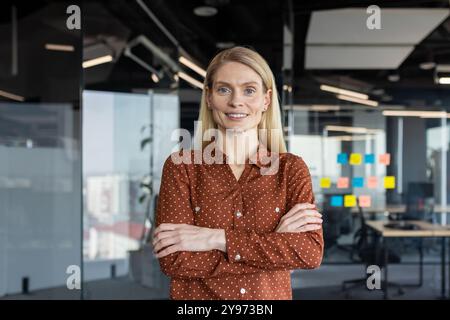 Smiling businesswoman standing with arms crossed in modern office. Professional demeanor exudes confidence and leadership. Ideal representation of success and empowerment in workplace. Stock Photo