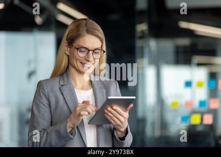 Businesswoman in gray suit interacting with tablet in office. Female professional using digital device with focus and precision. Elegant career woman embracing technology for work efficiency. Stock Photo