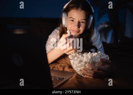 Teen girl wear headphones with long dark hair lying on bed watching interesting movie and eating popcorn on laptop at night Stock Photo