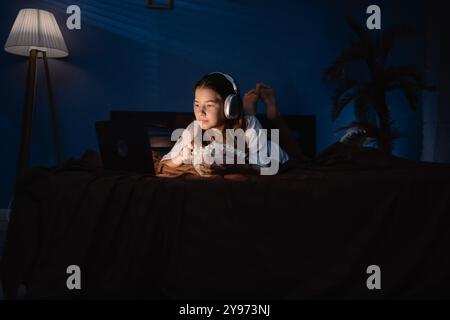 Teen girl wear headphones with long dark hair lying on bed watching interesting movie and eating popcorn on laptop at night Stock Photo