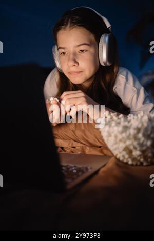 Teen girl wear headphones with long dark hair lying on bed watching interesting movie and eating popcorn at night Stock Photo