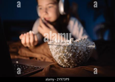 Teenage girl eats popcorn lying on bed at night watching interesting movie, popcorn in focus Stock Photo