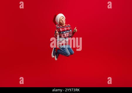 Full length photo of charming small boy jump show thumb up celebrate christmas dressed knitted sweater isolated on red color background Stock Photo