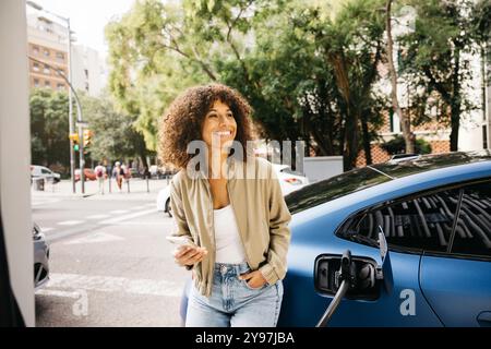 A woman is smiling as she stands next to her electric car at a charging station in a city environment. The scene captures modern transportation and ec Stock Photo