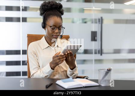 Professional woman engaged with tablet in office while wearing headset. Interacting with digital content, multitasking, remote communication. Efficient workspace enhancing productivity. Stock Photo