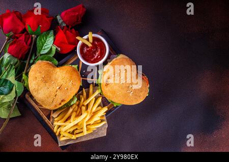 Valentine day Heart-shaped burgers. Two tasty cheeseburgers with french fries and beer bottles on dark table background. Idea for Valentine day dinner Stock Photo