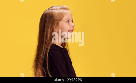 Side profile of little girl sticking her tongue out, smiling slightly against yellow studio background. Copy space. Ad Stock Photo