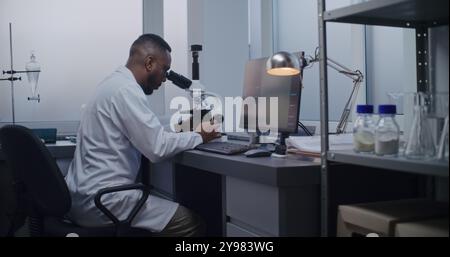Medical science laboratory: African American microbiologist looks under microscope, does analysis of test sample using high-tech equipment. PC screen shows image of cells or bacteria. Microbiology. Stock Photo