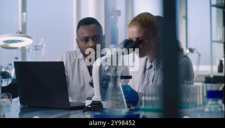 Multiethnic team of scientists working in medical laboratory. Female microbiologist examines drug samples under microscope, discusses research with male colleague entering results into computer. Stock Photo
