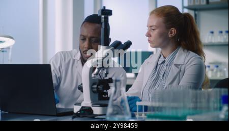 Multiethnic team of scientists working in medical laboratory. Female microbiologist examines drug samples under microscope, discusses research with male colleague entering results into computer. Stock Photo