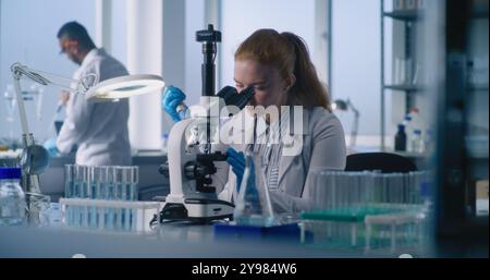 Young female microbiologist looking under microscope, adding new samples, studying and comparing results. In the background, male scientist works with samples. Advanced medical science laboratory. Stock Photo