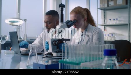 Advanced medical science laboratory: Two diverse scientists using laptop to develop new drug formula or vaccine, doing microscope analysis of samples from test tubes. Medicine and pharmaceuticals. Stock Photo