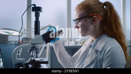 Female scientist or chemist wearing protective glasses to analyze and study new sample under microscope, uses laptop computer. In the background doing analysis. Modern medical science laboratory. Stock Photo